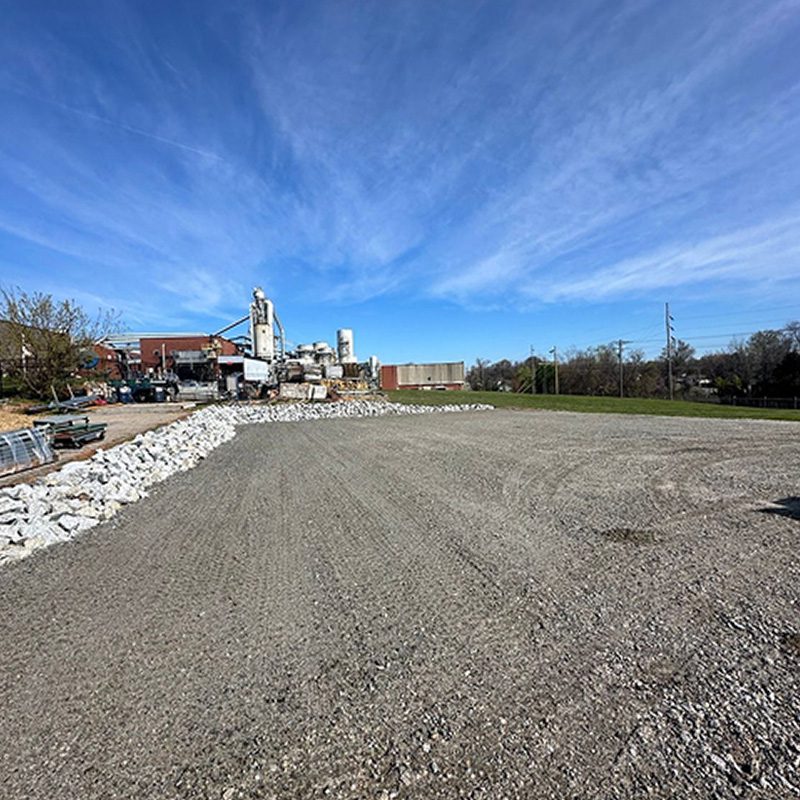 Industrial construction site with a gravel lot, rock piles along a low wall, and a factory building under a blue sky.