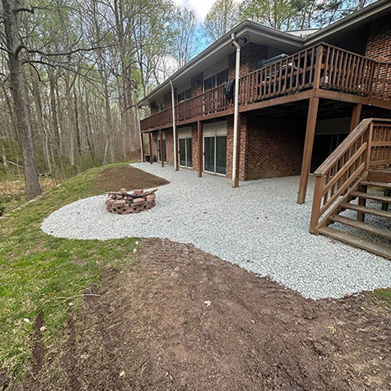 Backyard patio area under construction with a circular fire pit, light-gray gravel, and a wooden deck attached to a brick house.